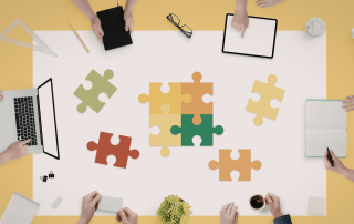 Immagine di mani di diverse persone riunite attorno a un tavolo di lavoro, come in un team: alcune sono su tastiere di computer, altre vicine a una tazza di caffè. Al centro del tavolo ci sono pezzi di puzzle sparsi che rappresentano il concetto di job crafting, simbolo di costruzione e personalizzazione del proprio lavoro.